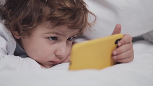 Little boy lying in the bed watching cartoon at modern yellow smartphone with interest.