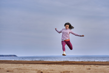 Obraz premium Young woman with black hair jumps while jogging. A woman is engaged in gymnastics in the spring morning on the sandy bank of a large river. Cloudy morning.