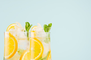 Cold oranges beverage with pieces fruit, ice cubes, mint on elegant soft light green background, closeup, top section, edge.