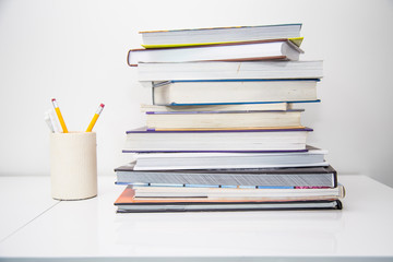 Home office desk with a high stack of colourful books in a pile and a container with pens and pencils.  Sitting on a white desk gives a light, bright and airy feeling about this busy home business.
