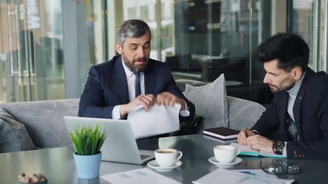 Angry businessman mature bearded man is tearing papers throwing at partner during tense talks in cafe. Argument, negotiations and communication concept.