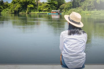 Foto op Canvas Alone Asian woman wear weave hat and white shirt with sitting on wooden terrace, she looking forward to river. © Angkana