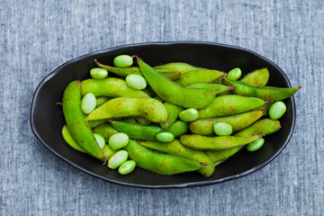 Fresh edamame green beans in black bowl. Grey textile background. Top view.