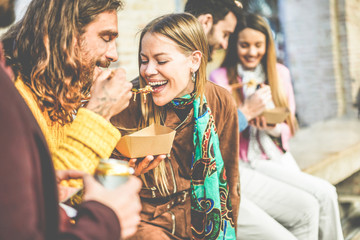 Happy friends eating street food outdoor - Young trendy people having fun together drinking and laughing around downtown streets - City lifestyle and party concept - Focus on blond girl face