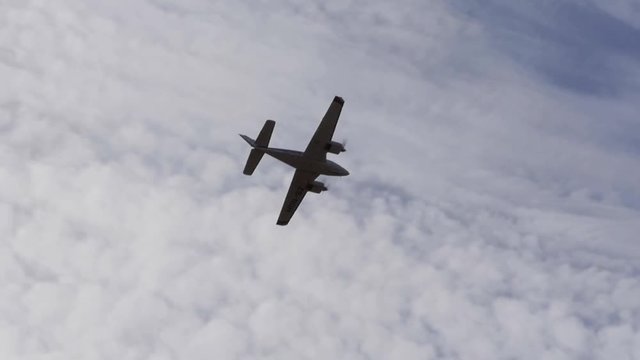 A twin prop light aircraft flies overhead silhouetted by the clouds