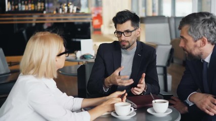 Bearded businessman in trendy suit is speaking with male and female partners in modern cafe discussing work. Communication, business people and lunch concept.