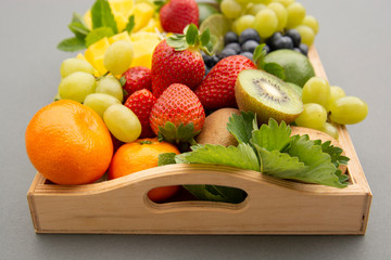 Various fresh fruits - mango, grapes, tangerine, lime, strawberry, kiwi, mint, on wooden tray and a gray background.