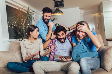 Four casual happy friends laughing shopping online together in a tablet sitting on a sofa in the living room at home