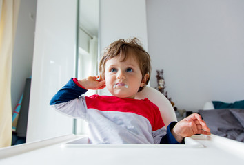 Little toddler boy sitting in a chair for feeding in a room in breakfast time