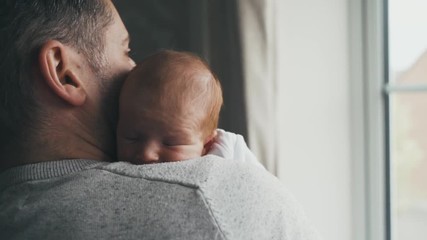 Sweet, tiny baby with thin red hair snuggles against daddy. Sunlight through a window shines on father and newborn baby as they cuddle. Dad and baby cheek to cheek in a peaceful moment. PUSH IN. - Powered by Adobe