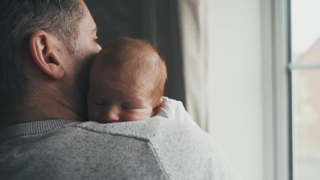 Sweet, tiny baby with thin red hair snuggles against daddy. Sunlight through a window shines on father and newborn baby as they cuddle. Dad and baby cheek to cheek in a peaceful moment. PUSH IN.