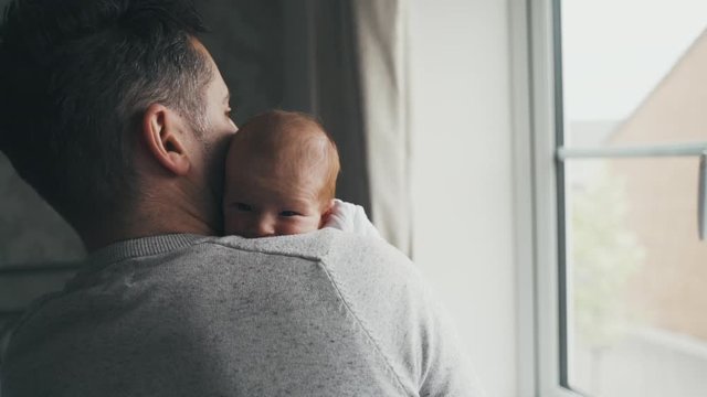 Dad holding baby near a bright window. Father comforts and cuddles infant while standing. Parenthood, fatherhood and love concept. Man with baby against his chest, head on shoulder. Slow motion.