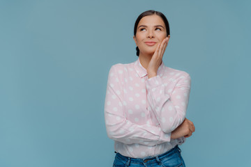 Portrait of cheerful satisfied brunette female touches cheek gently, looks above, dreams about something pleasant, wears polka dot shirt and jeans, stands against blue background with copy space