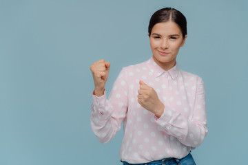 I am confident, I will win. Beautiful serious brunette woman keeps hands clenched in fists, ready to defend herself, dressed in polka dot shirt, smiles contendely, gestures over blue background.