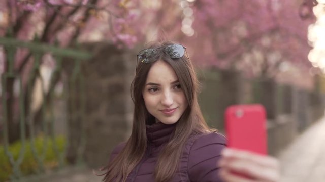 Young Caucasian Female With Sunglasses on Head Taking Selfies With Smartphone Under Spring Tree in The Park. Light Camera Movement
