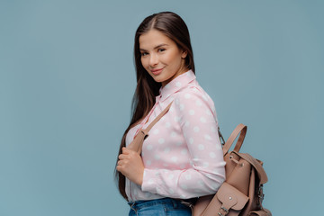 Fashionable delighted schoolgirl carries rucksack, returns from school, going to have extra classes, wears polka dot shirt, looks happily at camera, isolated over blue background. Woman with backpack