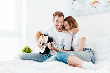 Smiling man sitting on bed with wife and playing guitar