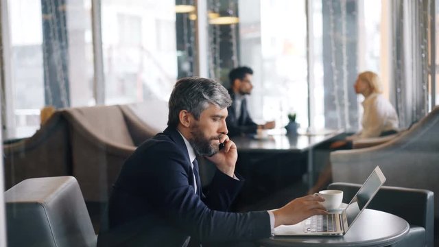 Entrepreneur handsome bearded man in talking on mobile phone and using laptop working in cafe alone. Modern technology, communication and work concept.