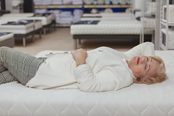 Relaxed senior woman trying orthopedic mattress on sale at furniture store. happy elderly female customer smiling with her eyes closed, lying on a new bed at home department shop