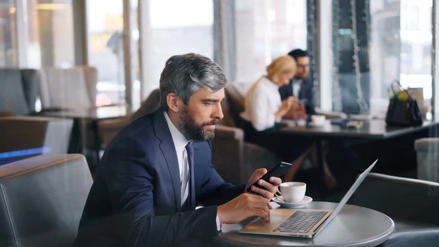 Mature businessman bearded grey-haired man is using laptop and smartphone during business lunch in cafe. Modern technology and entrepreneurship concept.