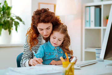 Red-haired mom helping her little girl holding pen and writing