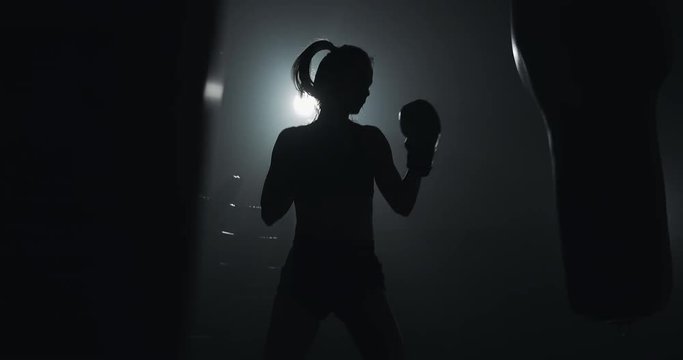 Silhouette of young woman training with a punching bag in a boxing club. Slow motion. Darkness background
