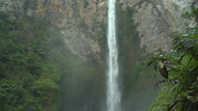 Vertical pan footage of a woman walking across a wooden bridge at the base of Sipiso Piso waterfall in North Sumatra, Indonesia.
