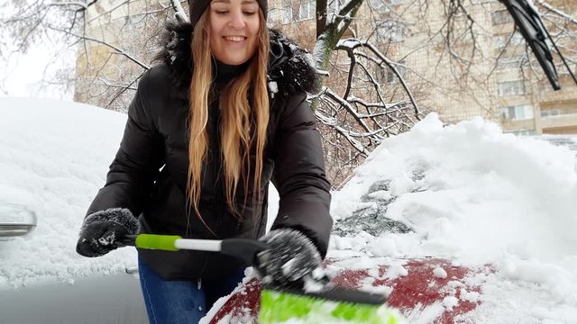 4k video of beautiful smiling young woman cleaning car windshield from snow with brush