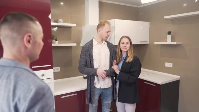 Young Caucasian Married Couple Standing in Kitchen of New Apartment Property and They Talking With Male Realtor Real Estate Broker. Light Camera Movement