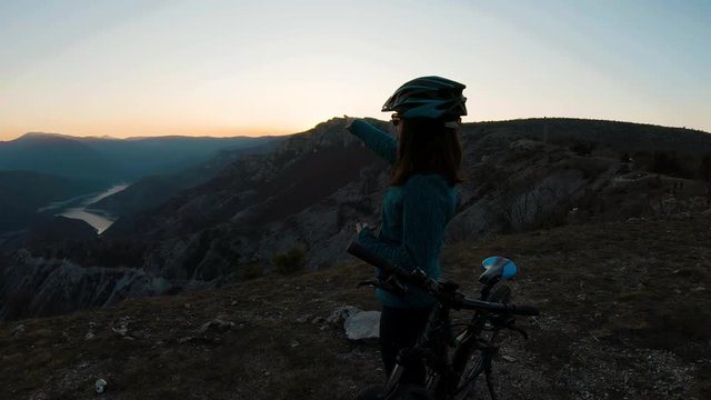 Young biker girl explaning and pointing to a caynon lake from a top of a mountain at a sunset
