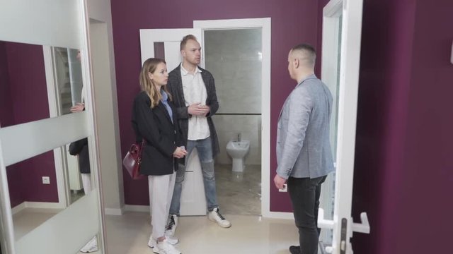Young couple is looking around a new flat. There is a realtor next to them who is showing them the rooms and answering on their questions.