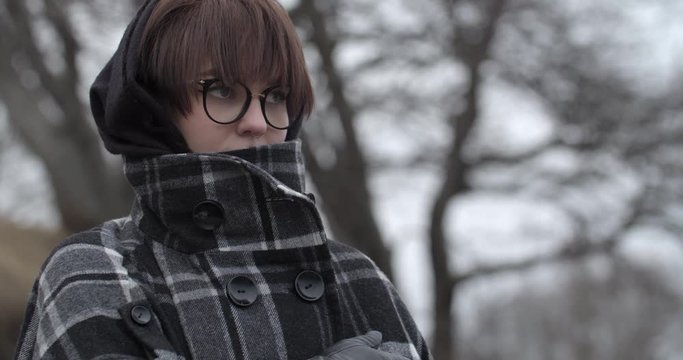Beautiful young woman standing infront of the camera and gazing into the distance while wrapping herself up in a jacket to keep warm, depicted through a close up shot that focuses on the woman's face.