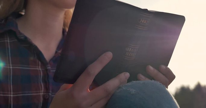 Close up of a girl reading The Bible in a forest on a sunny day. Lens flare showing up behind.