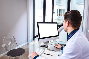 Young doctor working with computer with blank screen on the desk of a modern medical clinic.