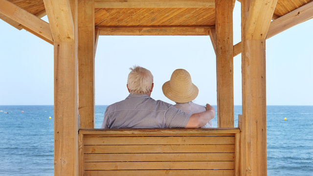Senior couple, woman with hat and man with white silver hair sitting under a wooden canopy on the promenade and looking at the ocean in a romantic embrace in El Medano, Tenerife, Canary Islands, Spain
