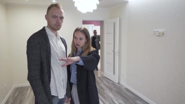 Young Married Couple Standing In Empty Room in New Apartment Real Estate Property and Talking about Future Arragements While Realtor Standing in Background. Light Camera Movement