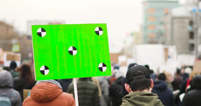 Revolution and demonstration in city during day. Man in jacket and cap holds poster with green screen and walking the street with crowd on city rally, back view.