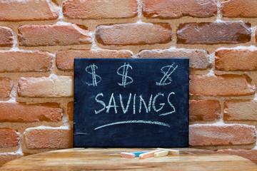 Black board with the word "Savings" drown by hand on wooden table on brick wall background.