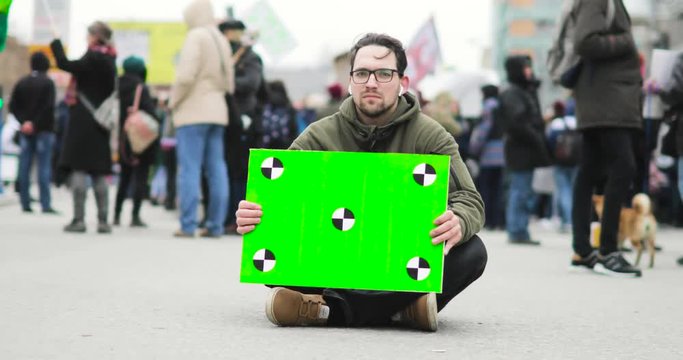 Poster with green screen in man's hands who is sitting on the street during demonstration in city with crowd on background.