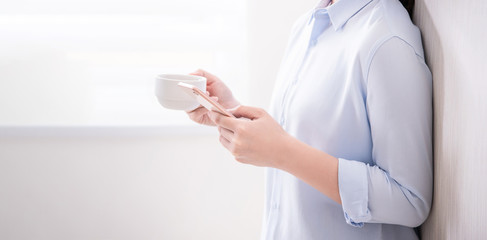 Obraz premium Side view of asia young business woman in blue shirt standing lying on the wall beside window drinking and using smart phone, copy space, close up