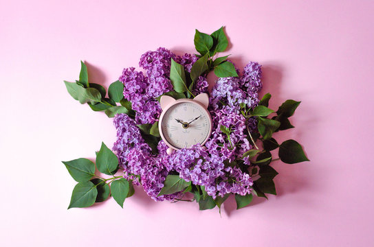 Pink alarm clock in the form of a cat lying on a bouquet of lilacs.