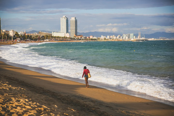 Girl with a bag walking on the beach on sunny day. Stylish hipster near the waves on the sea. Holiday travel concept. Barcelona Spain.