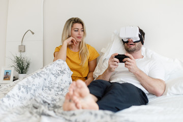 Joyful man wearing virtual reality headset for playing games ignoring his wife in the bed....