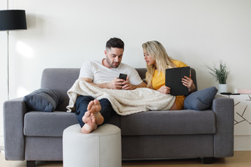 Young casual relaxed couple at home using smartphone and tablet sitting on a sofa. Man and woman with digital online connected devices. Generation Y relationship with technology concept.