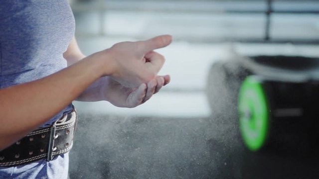 Slow motion of unrecognizable female athlete with weightlifting belt on her waist clapping hands applying chalk powder before weight training