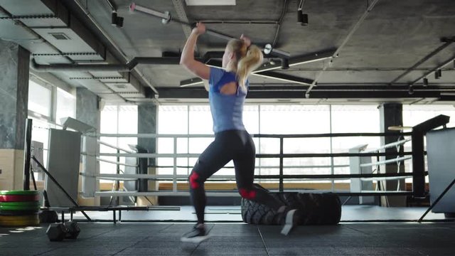 Three quarter back view of blonde female athlete lifting barbell and doing clean and jerk exercise during workout in gym