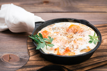 Homemade breakfast of shakshuka fried eggs with tomatoes and herbs in a pan on a wooden table