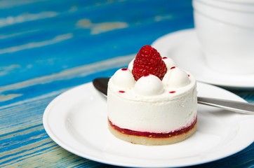 White airy cake with raspberry layer and raspberry berry lies on a white round plate next to a white cup, which stands on a blue table