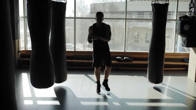 Tilt up of professional young boxer doing exercise with jump rope among punching bags in dark gym