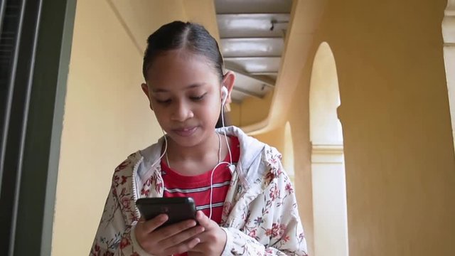 Cute girl listening music from mobile phone with earphones while walking in corridor of chino portuguese style building.